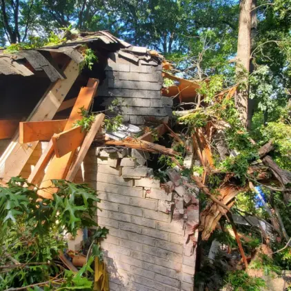 Storm-damaged house with a collapsed roof and exposed brick chimney surrounded by fallen tree branches.