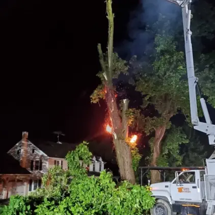 Tree branches sparking and burning at night near a house roof, with an aerial lift boom positioned alongside.