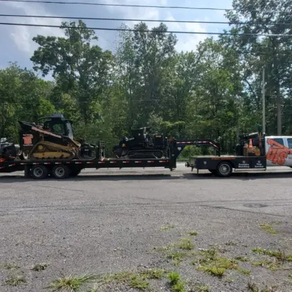 A pickup truck towing a trailer loaded with two compact track loaders parked along a tree-lined road.
