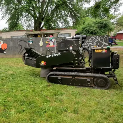 Compact tracked stump grinder parked on a lawn beside a cut tree stump and a decorated backyard fence.