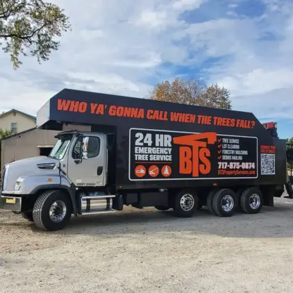 Tree service dump truck advertising 24-hour emergency service, parked on a gravel lot with another boom truck visible behind it.