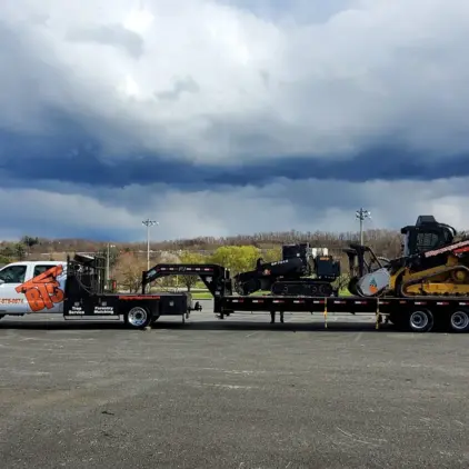Pickup truck towing a trailer loaded with a stump grinder and skid steer under a dark, stormy sky.