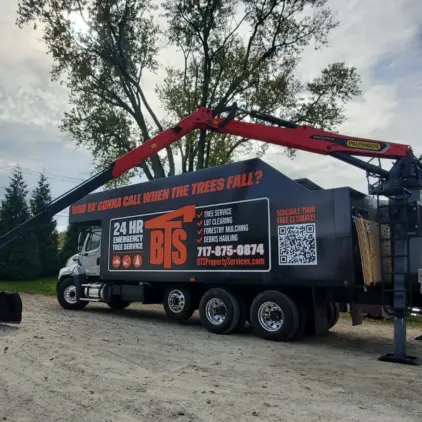 Tree service truck with a red knuckle-boom crane and grapple bucket parked on a gravel road beside evergreen trees.