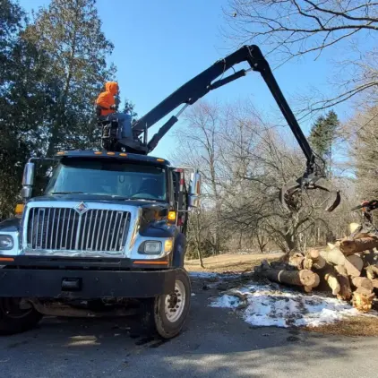 A grapple loader mounted on a truck lifts logs from a woodpile beside a brick house in a leafless winter yard.