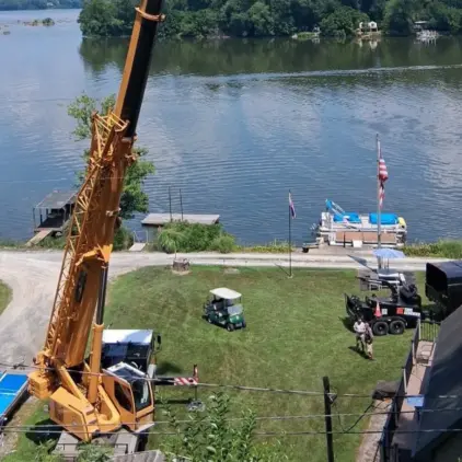Aerial view of a mobile crane's boom reaching toward a lakefront yard with a dock, boats, and a golf cart parked nearby.