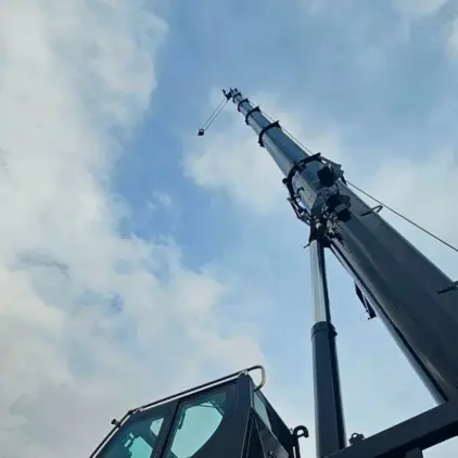 Looking up at a fully extended crane boom and rigging hook against a cloudy sky.