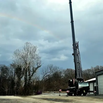A large hydraulic crane truck with outriggers extended sits outside a metal storage building near bare winter trees, with a worker standing nearby in high-visibility clothing.