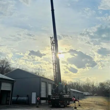 A mobile crane with its boom extended skyward and outriggers deployed is set up outside a row of metal storage buildings at dusk.