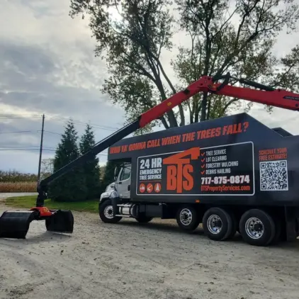 A flatbed truck fitted with a grapple loader arm and covered in tree-service and debris-removal advertising is parked on a gravel driveway.
