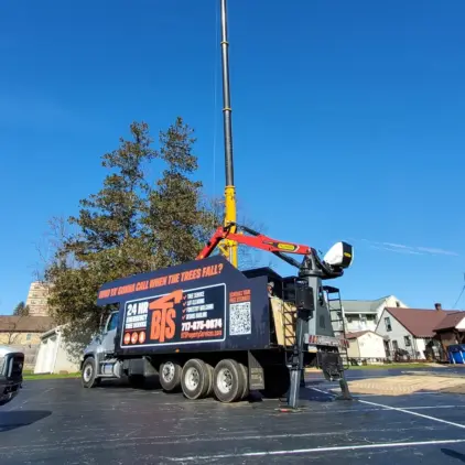 A heavy tree-service truck with a hydraulic crane arm parked in a lot, signage advertising 24-hour emergency tree service.