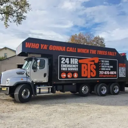 A large tree-service truck with an enclosed cargo box and crane arm parked outdoors, signage advertising 24-hour emergency tree service.